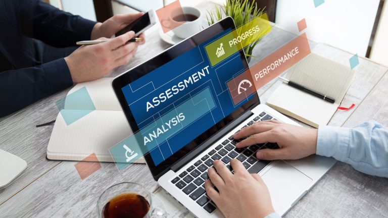 Deux personnes travaillent sur un ordinateur portable affichant les mots "assessment", "progress", "analysis" et "performance".